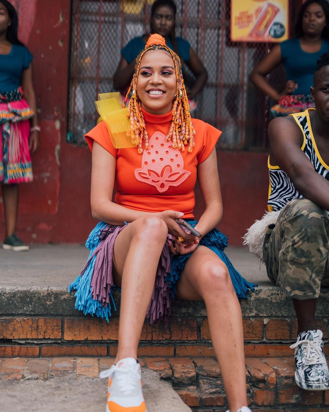 Sho Madjozi in pompom skirt and orange top with orange hair and beads